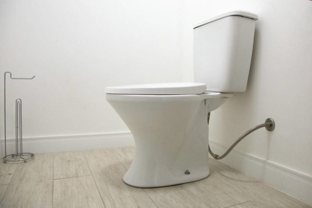White closed toilet with a metal toilet paper holder on a light wood floor against a white wall.